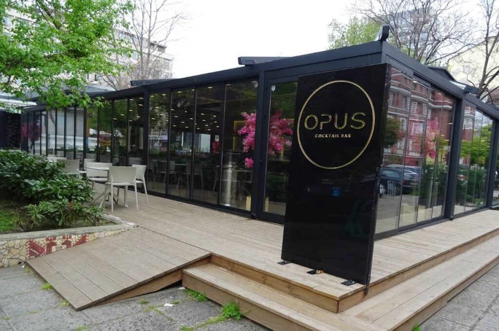 Външната част на бар Opus с изглед към стъклените врати и голяма табела с името на бара.